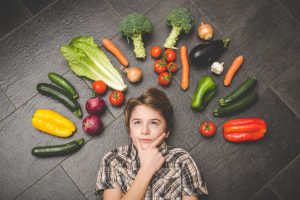 Vegane Kinderernährung: Junge nachdenklich auf dem Boden zwischen viel Gemüse